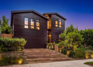Modern black house with large windows and wooden deck at dusk.